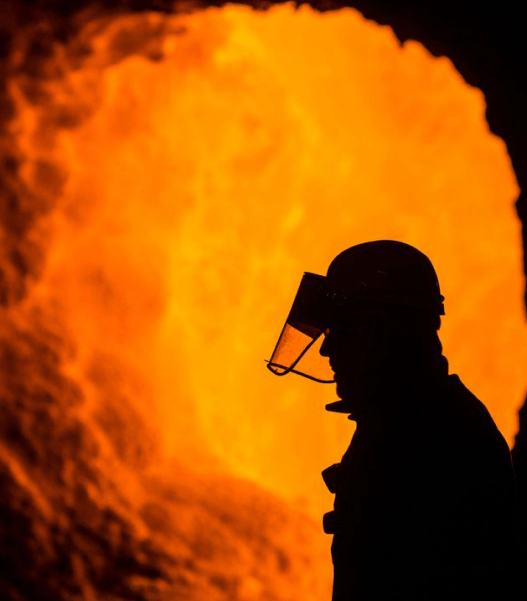 Silhouette of a metallurgical plant worker wearing a protective face shield in front of a roaring industrial blast furnace